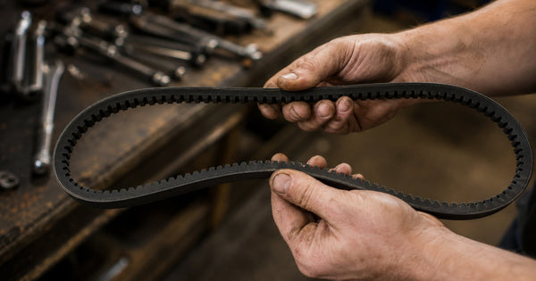 Industrial worker's two hands holding a black cogged V-belt open in a loop clearly showing the notched trapezoidal cog profile on the inner face with workshop tools softly blurred in the background