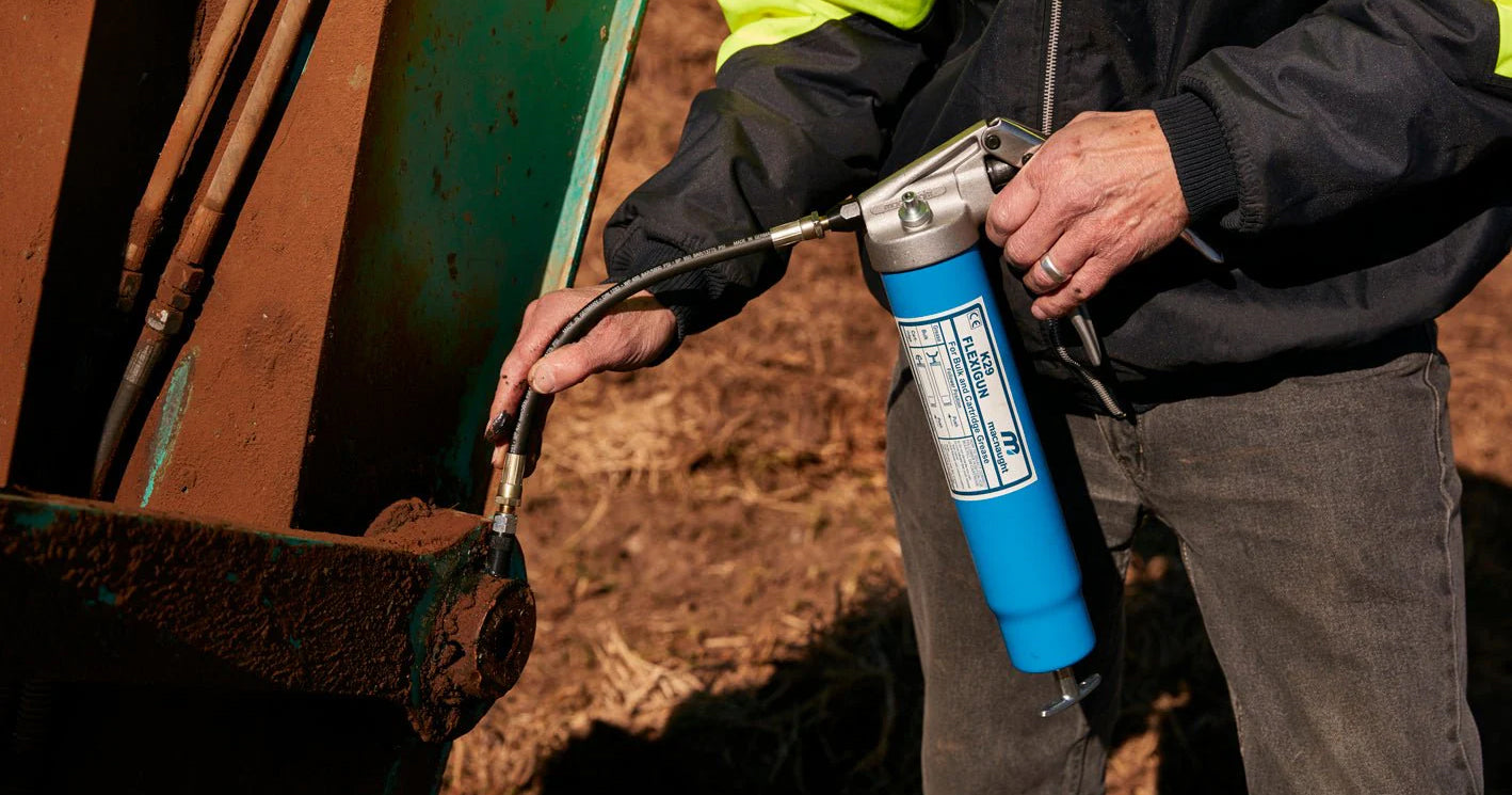 Maintenance worker in a high-vis jacket using a Macnaught K29 Flexigun lever-action grease gun with flexible hose to reach a recessed grease nipple on a heavy excavator or agricultural machine outdoors
