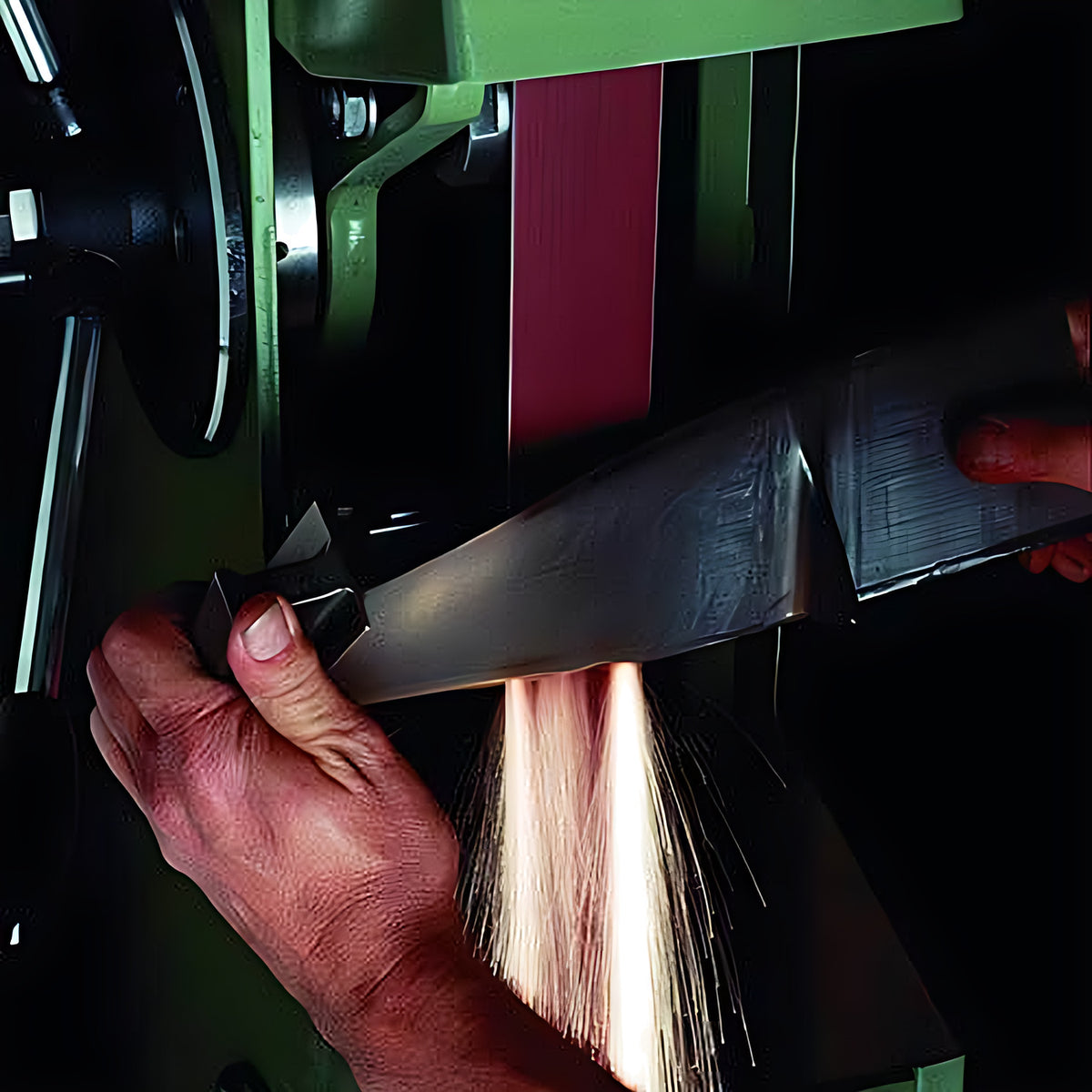 Pferd maroon aluminium oxide linishing belt 100 x 2440 mm on a belt grinder, hand holding a knife blade to the belt, sparks below